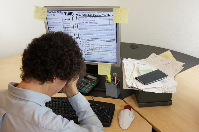A stressed taxpayer hold their head as they work on a desktop computer showing a IRS 1040 return with a nearby calculator, checkbook, and a stack of receipts