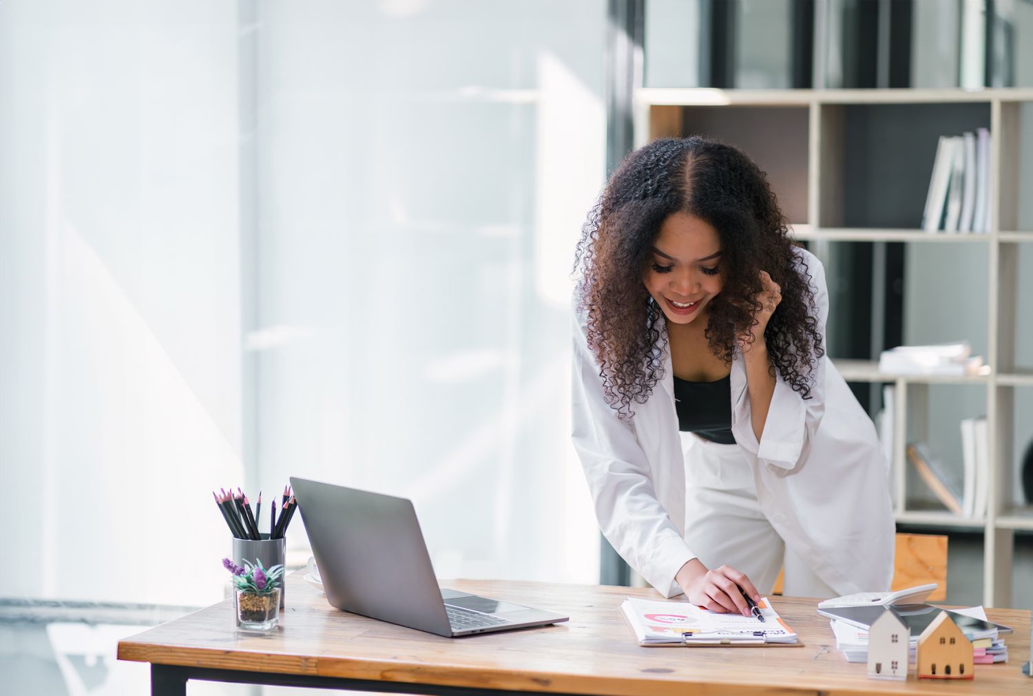 Smiling woman leaning over a desk while talking on a phone and writing on a notepad.