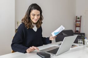A happy woman uses a calculator and laptop at a desk.