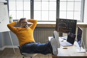Person sitting with feet up at a desk, two monitors showing graphs and data, relaxed posture in an office setting