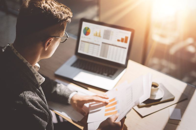 Close-up of a young man holding financial papers in his hand looking at his laptop that contains financial information