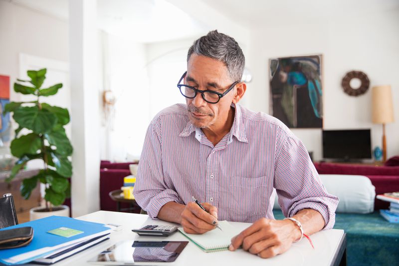A man with short gray hair and glasses sits at a desk in a bright, cozy room. He is focused on writing in a small notepad, with a calculator, tablet, and papers spread out in front of him. The background features a potted plant, artwork, and comfortable furnishings.