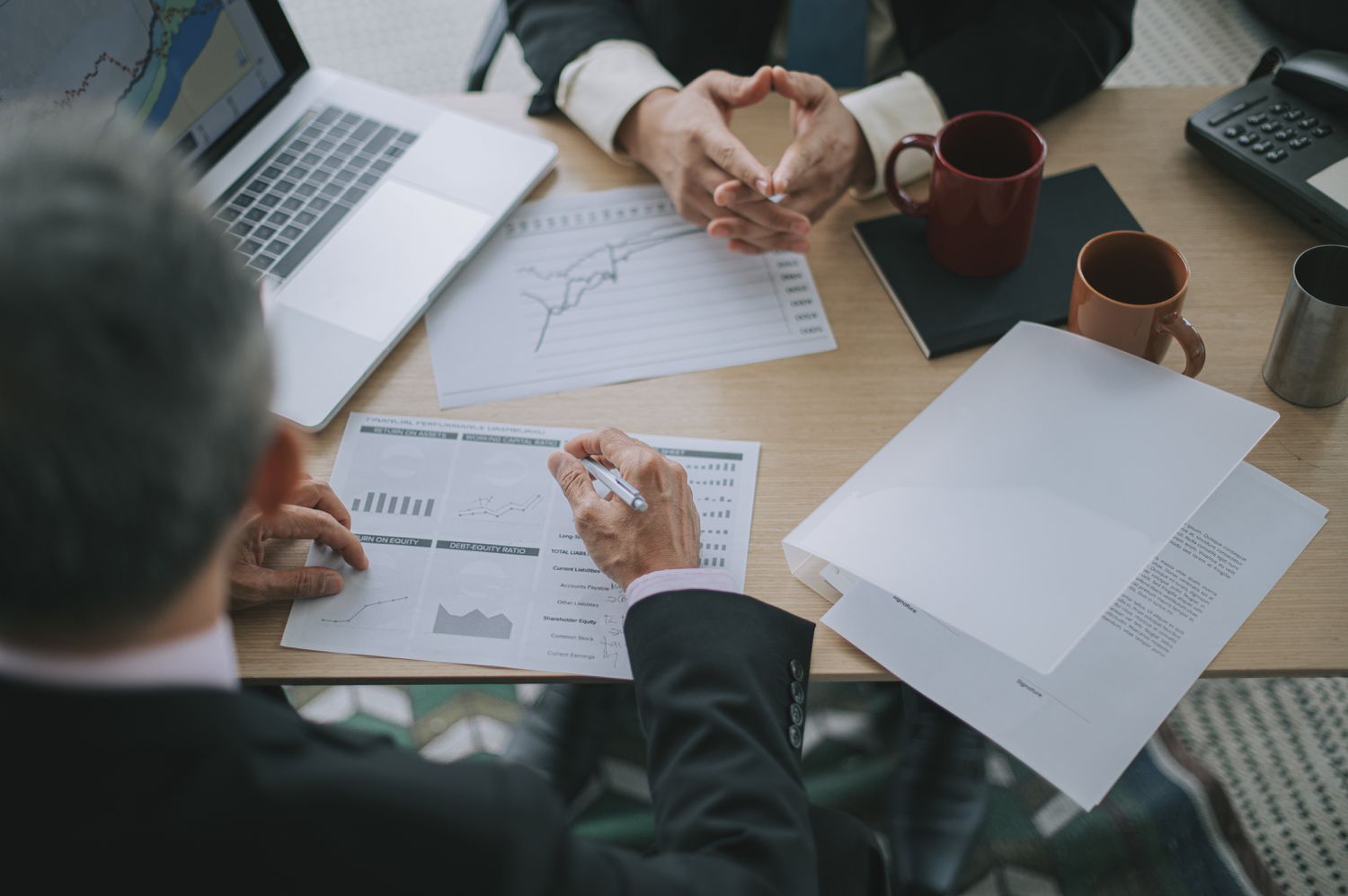Two managers share a desk, laptop, and financial reports to analyze the same-store sales figures for the previous quarter.