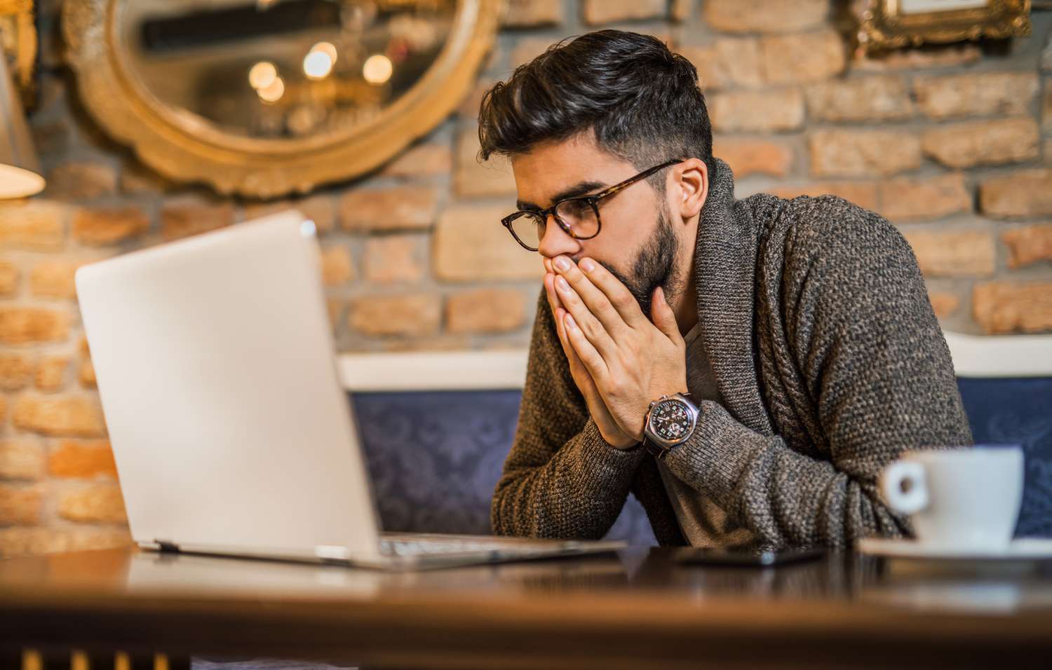 A person looks alarmed and covers their mouth with their hands as the read world events and economic news on laptop.