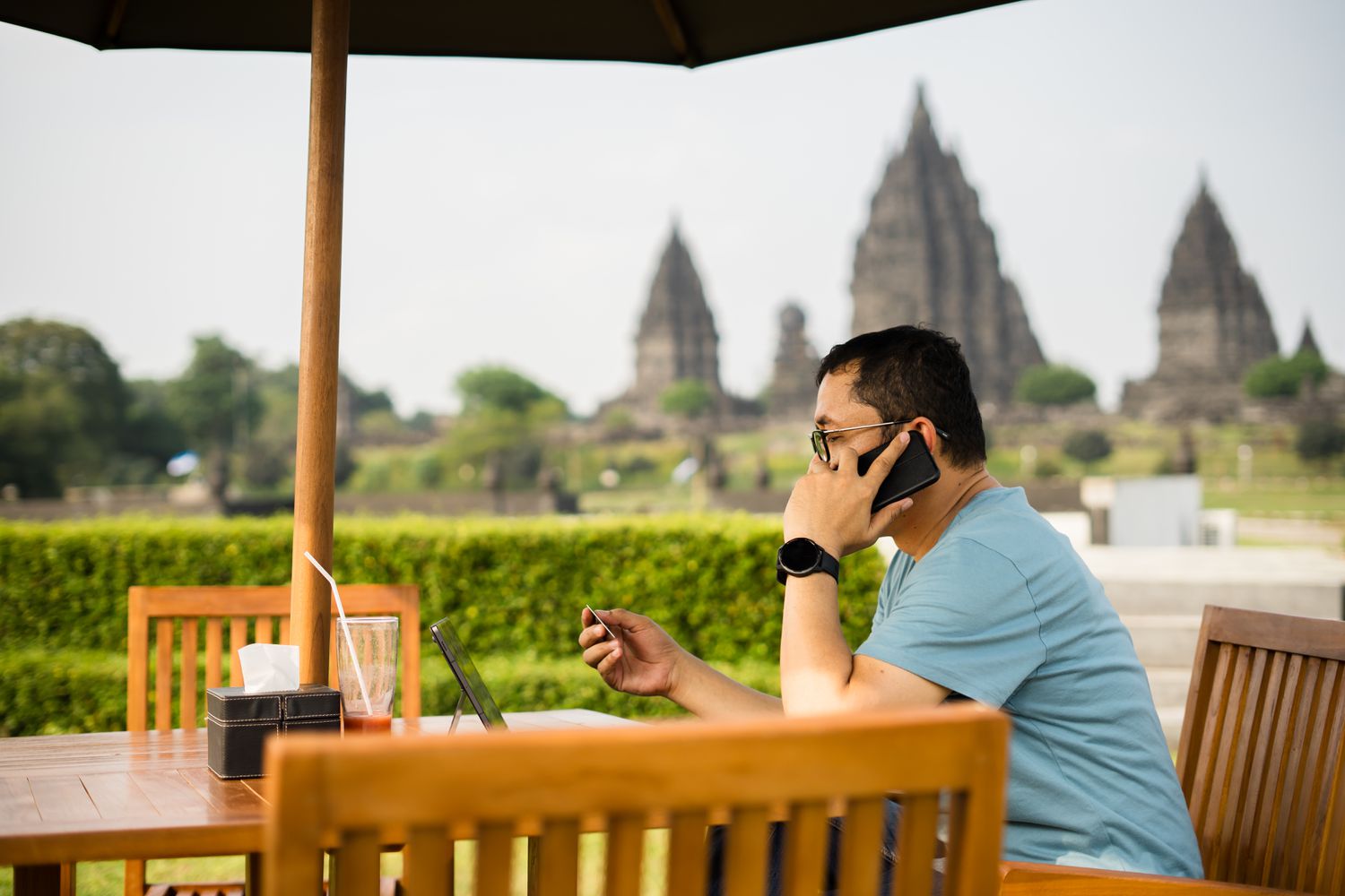A man sitting outdoors at a restaurant table using a cell phone with the Prambanan temple in the background.