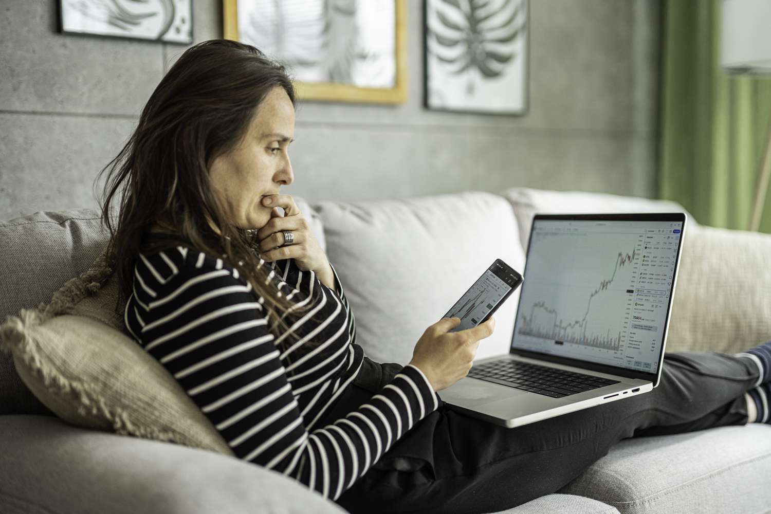 Woman sitting on a couch reviewing stock charts on a mobile and laptop