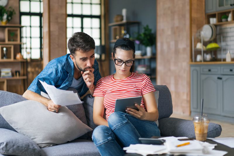 Woman on couch when man behind her with paperwork. Both are looking at an iPad with several papers, calculator and a coffee on the table in front of them.