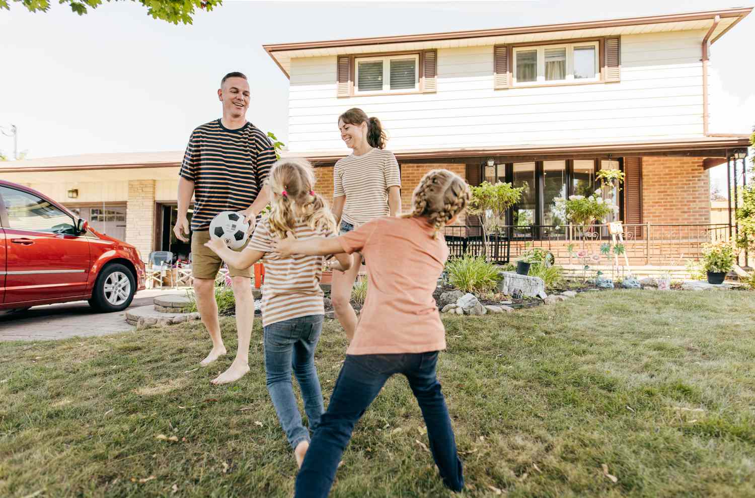 A family playing soccer in front of their house with a red car park in their driveway.