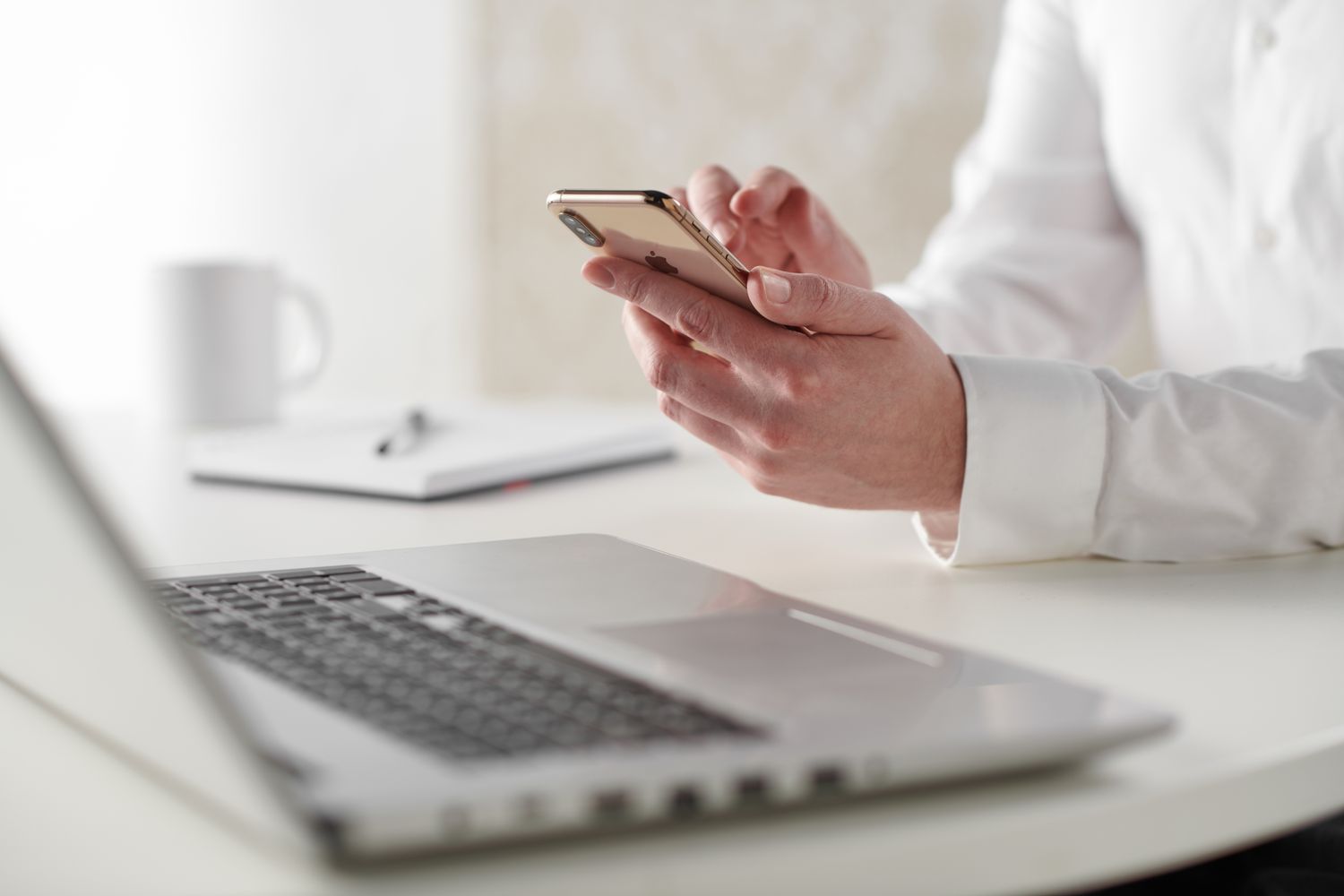 Close up shot of a person's hands holding a smartphone, with a lap top on the desk.