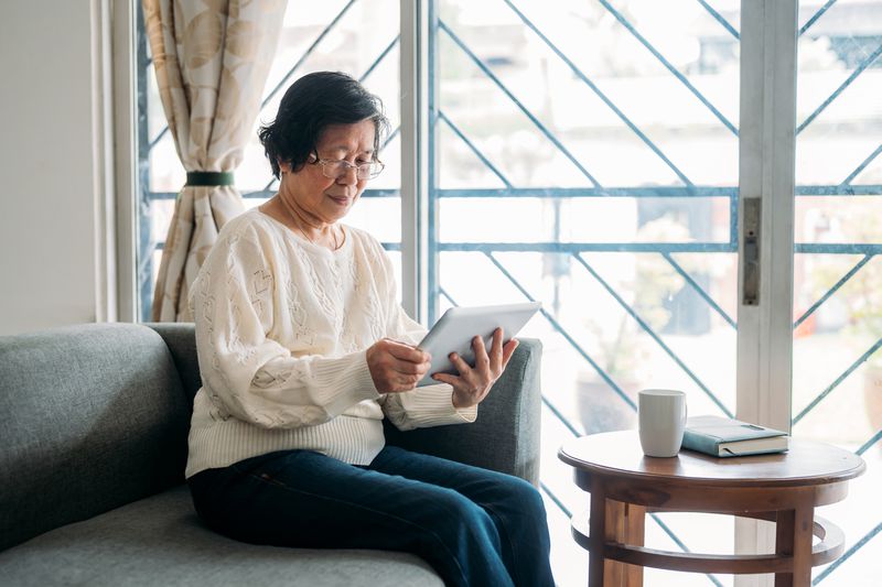 Woman in her 70s at home using a tablet. She is sitting on a grey sofa in front of a window in the living room.