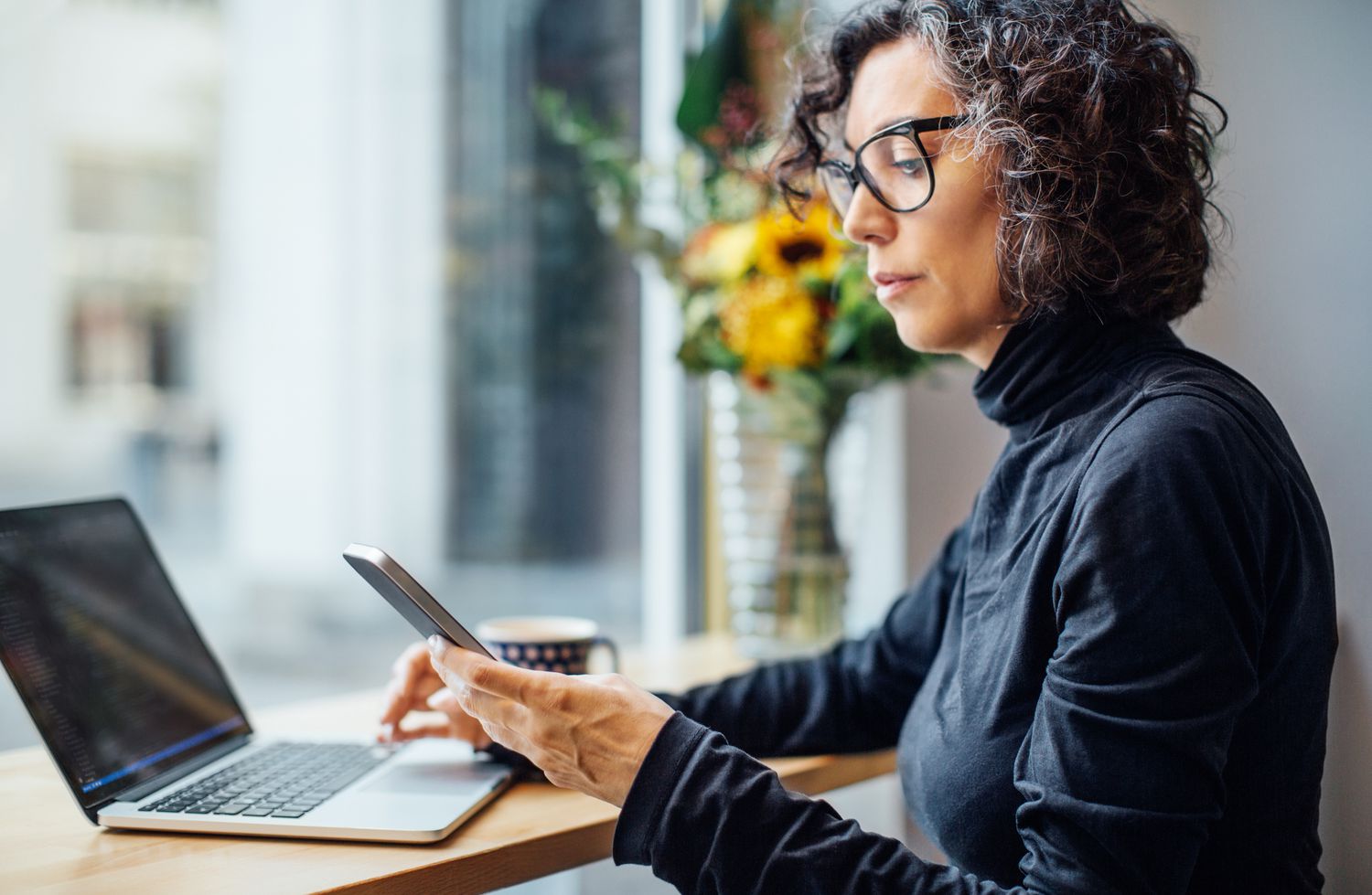 Mature woman sitting at cafe looking at her cell phone while working on laptop computer.