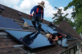 Two workers install solar panels on the roof of a home next to trees and a blue sky.