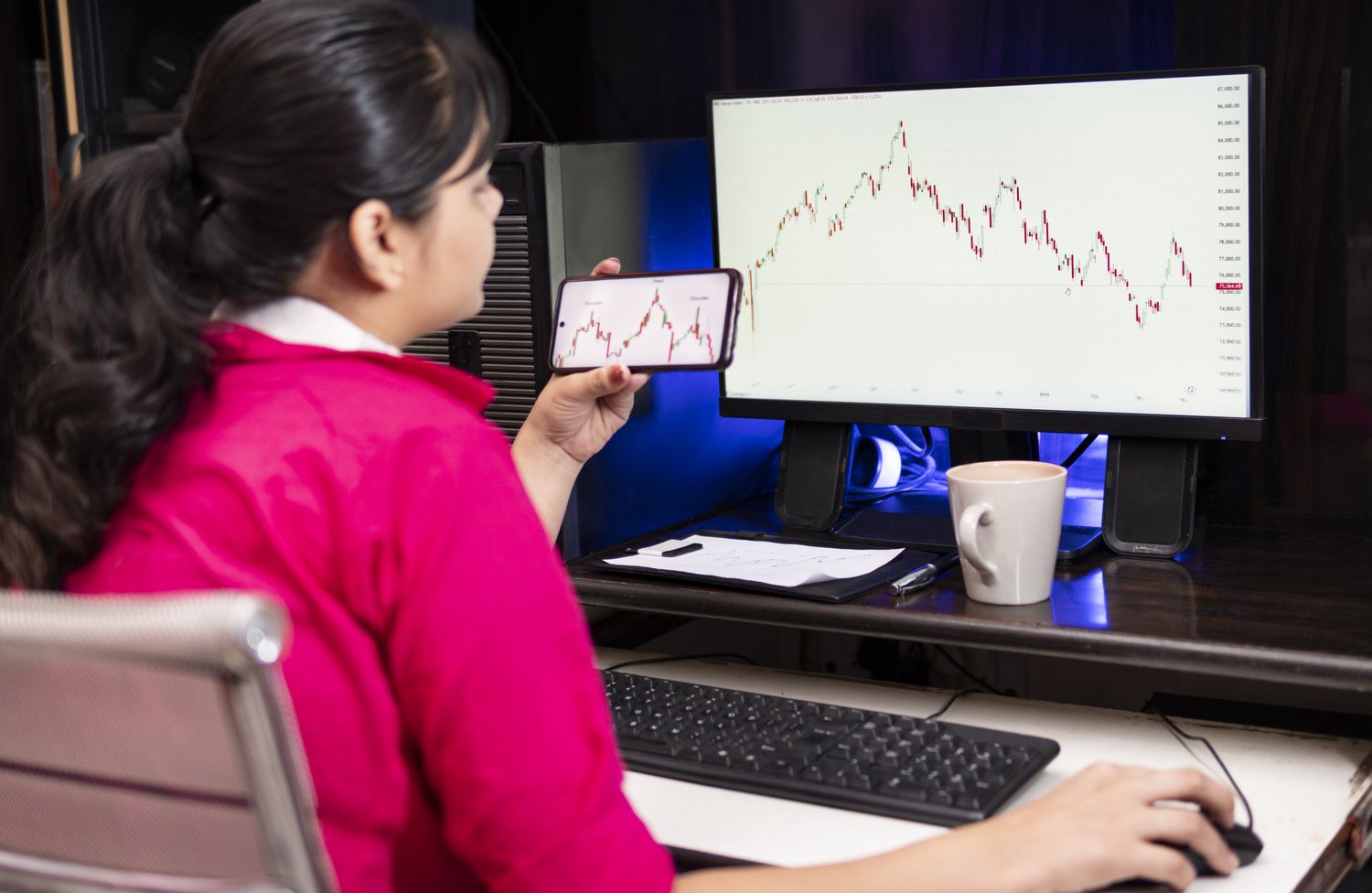 Young woman looking at charts on a monitor and a cell phone.