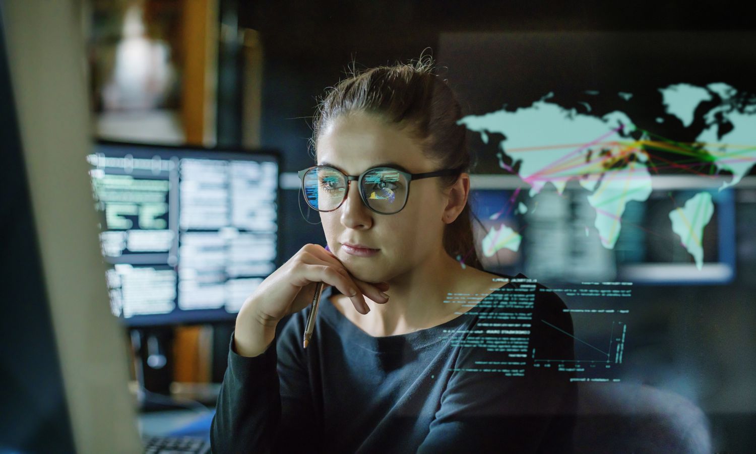 Stock image of a young person, wearing glasses, surrounded by computer monitors in a dark office. In front of her there is a see-through displaying showing a map of the world with some data.