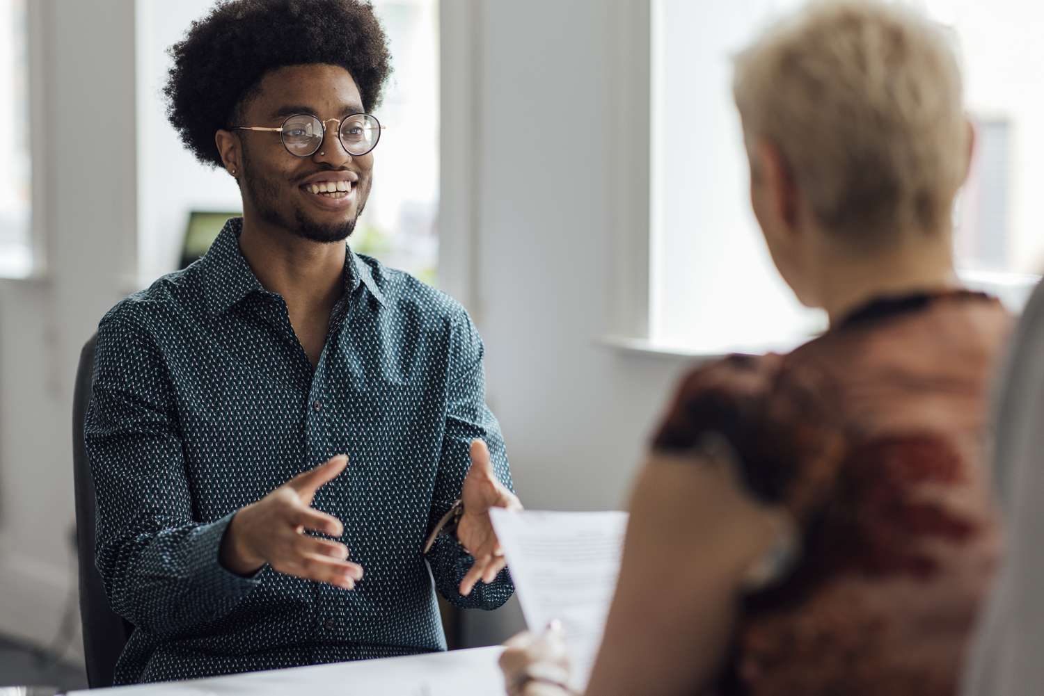 Over the shoulder view of a young male employee engaging in a focused discussion with a mature female manager at the managers desk in an office.