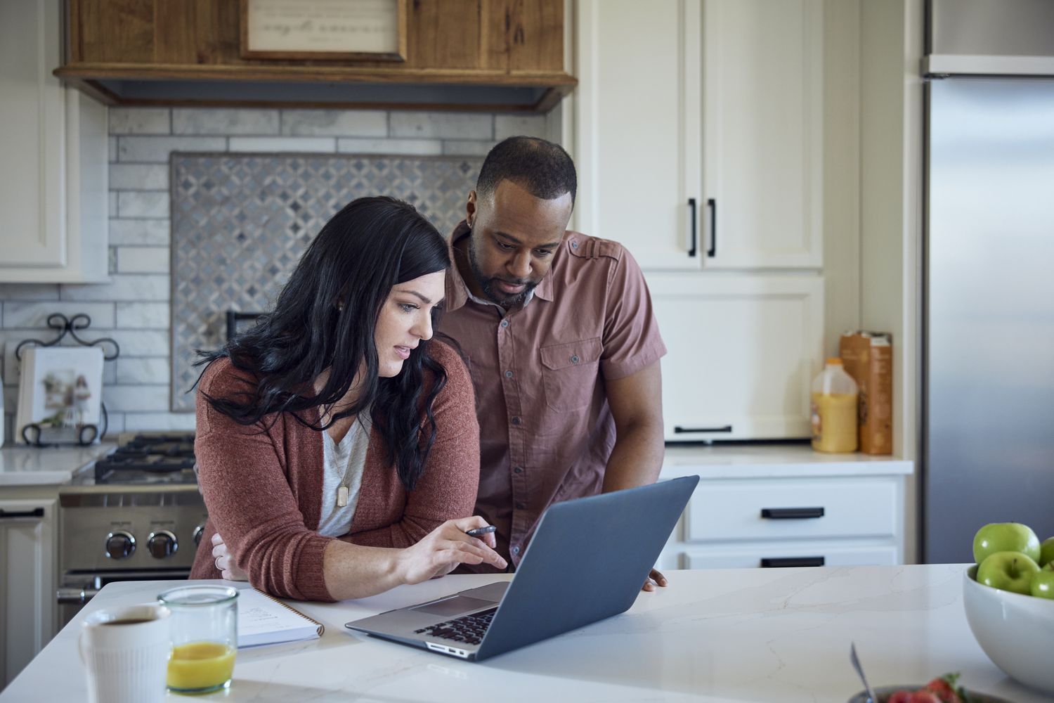 Woman and man sit at kitchen counter with breakfast working with pen, paper and laptop.