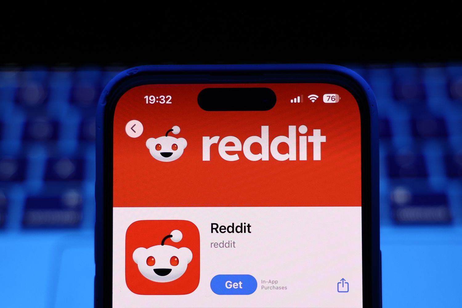 The App Store page for Reddit is seen on a phone screen, with a dark blue laptop keyboard in the background.