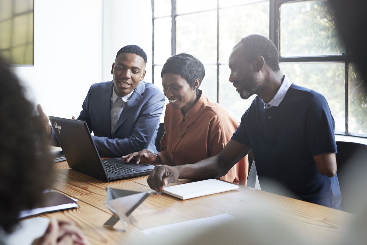 three colleagues in an office sitting at a table looking at a laptop and smiling