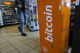A man walks past an ATM machine in a gas station. The ATM machine has the bitcoin logo on it.