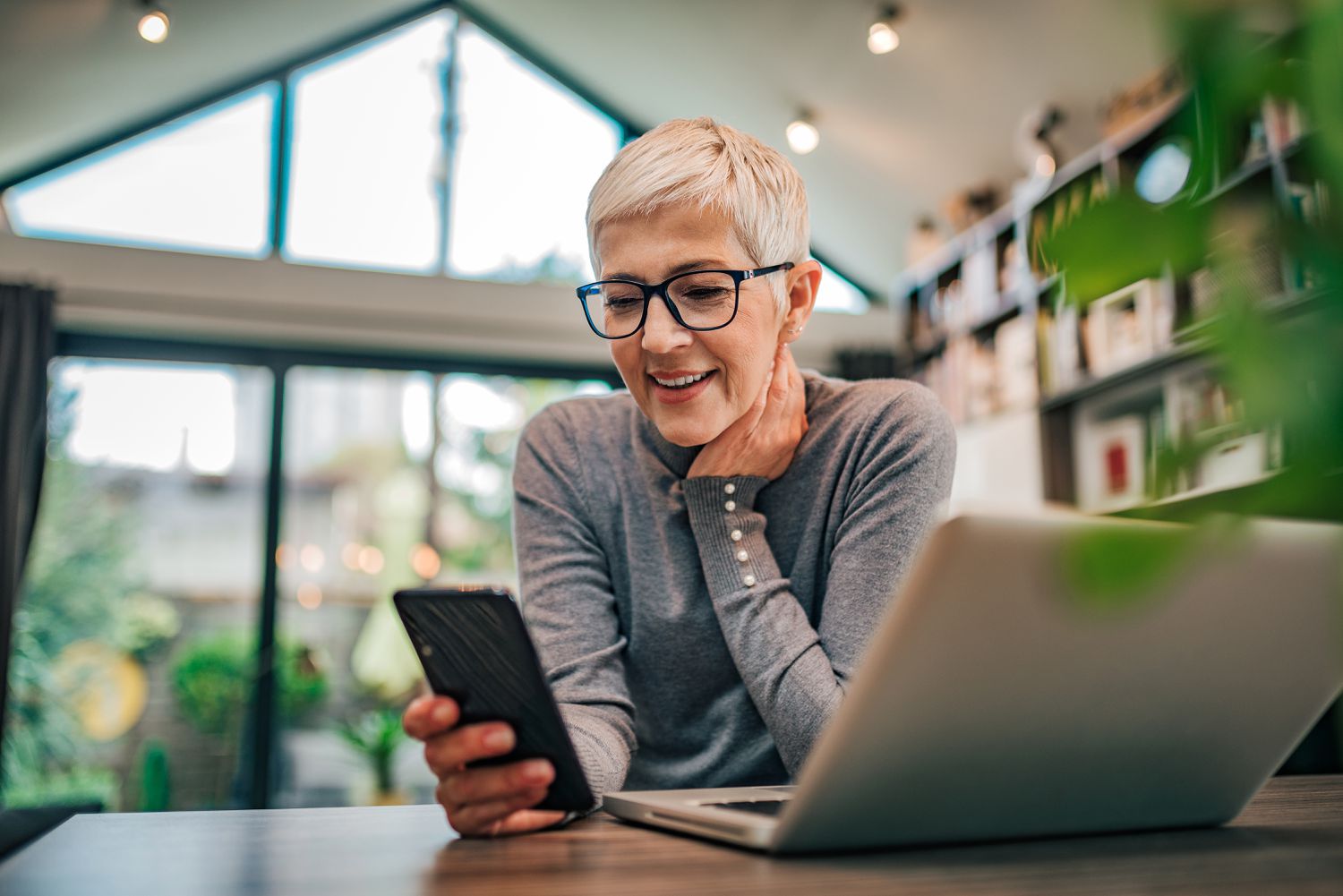 Older woman looking happily at her smartphone with a laptop also open in front of her