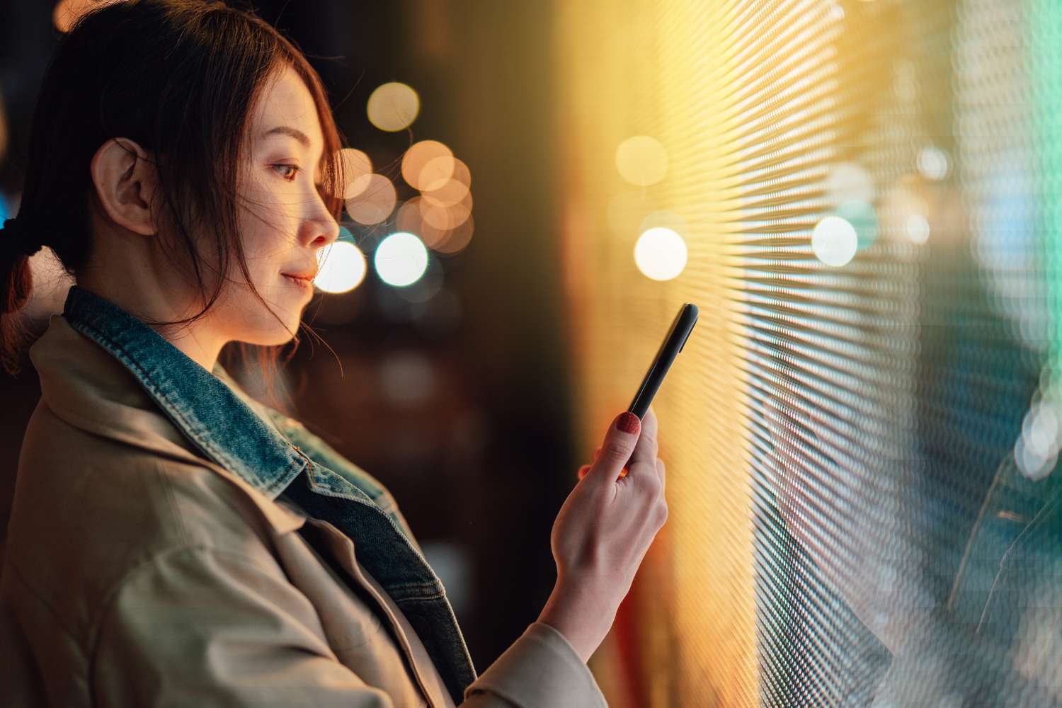 A person looks at a smartphone while illuminated by a digital display at night.