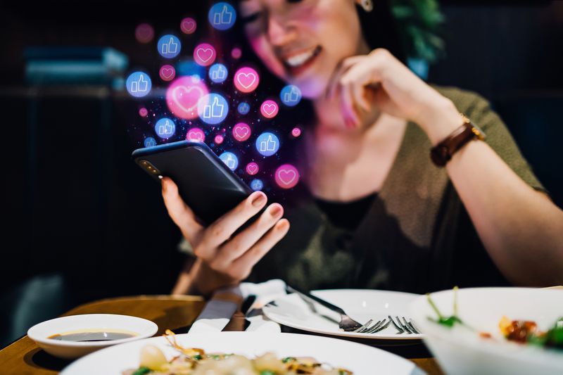 Young woman sitting at a restaurant looking at her phone with Facebook graphics of hearts and thumb likes coming out of it