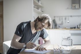 Man in casual clothing sitting at home writing on papers on the kitchen table