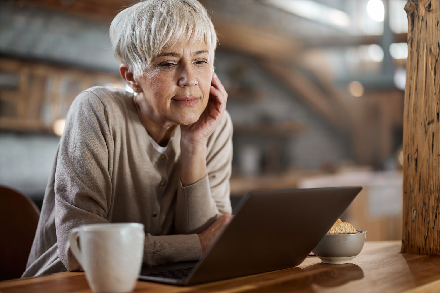 A woman with short white hair sits at a table with a laptop, a and a bowl of snacks