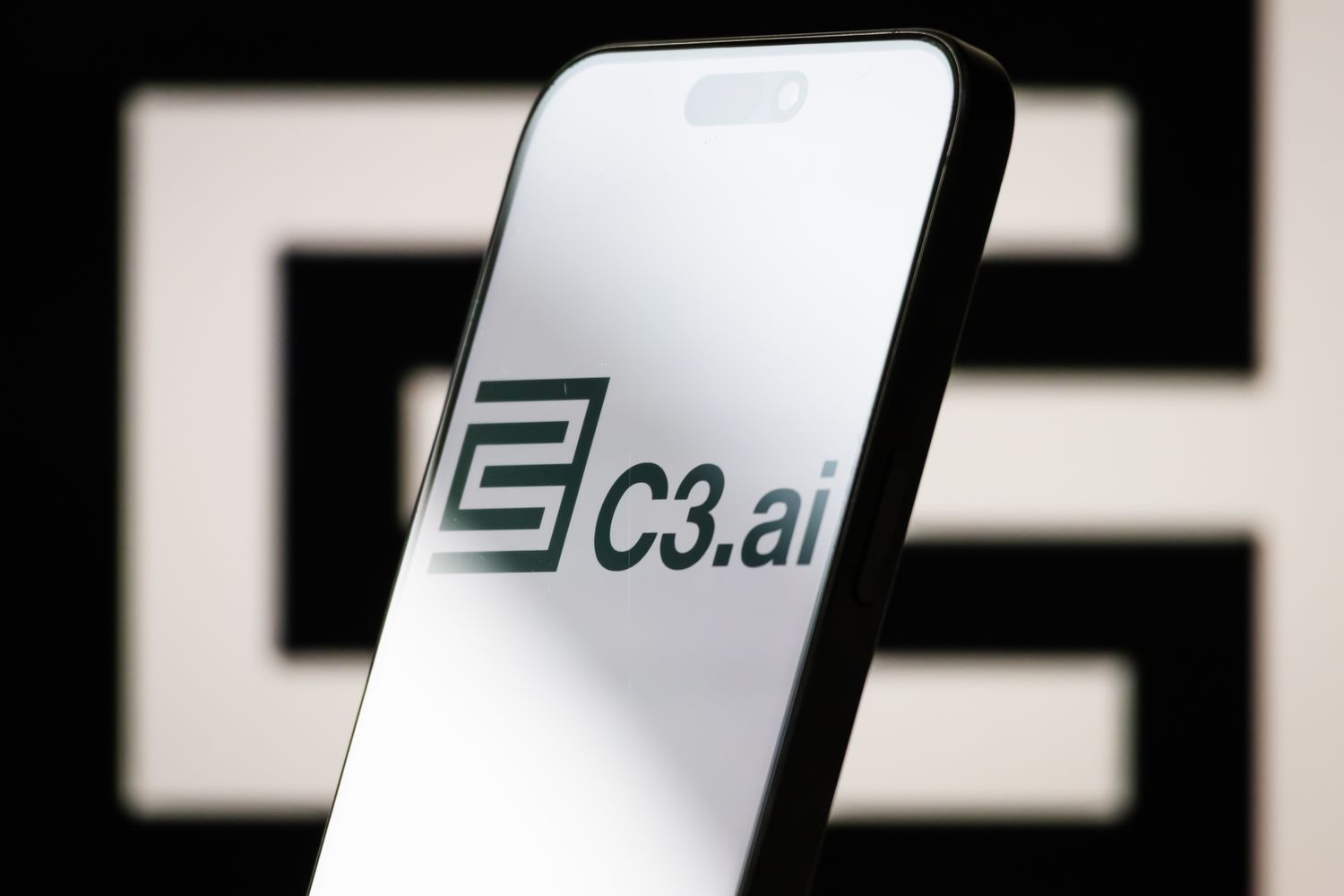 The logo of C3.ai is displayed on a smartphone screen, with the company's geometric black-and-white branding visible in the background
