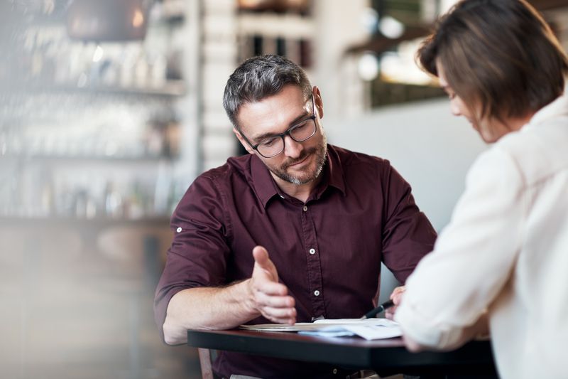Two adults sitting at a table discuss documents.