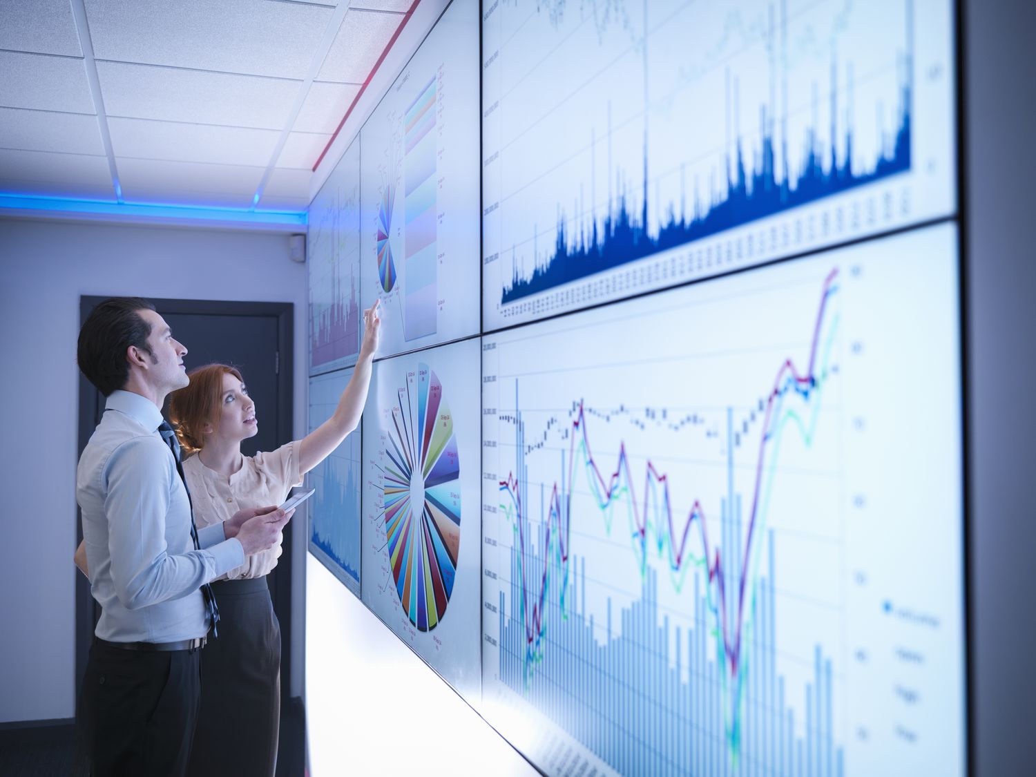 Two analysts study financial charts and graphs on several large screens before a company meeting.