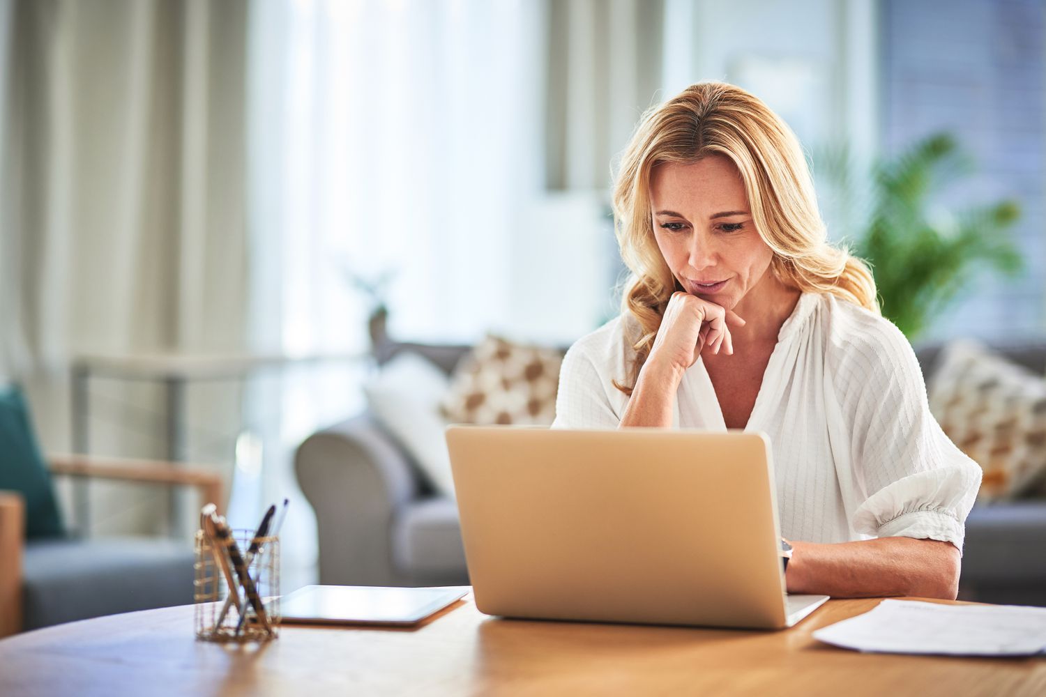 A woman in a white shirt sits at a desk at home with a laptop