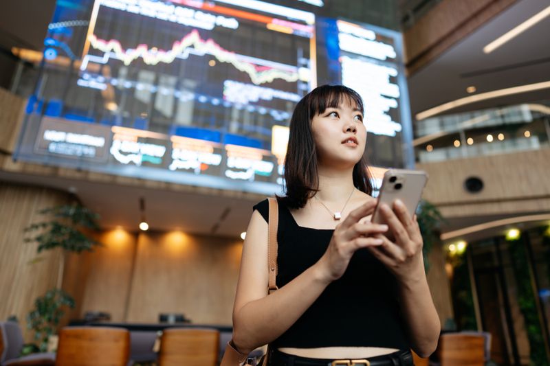 Young woman sanding with her back to a large screen showing market data and charts.