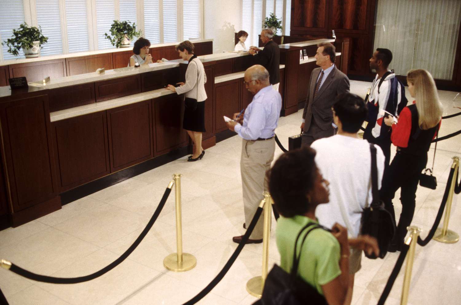 Several customers queue in the lobby of a large bank while tellers assist other customers at counters.