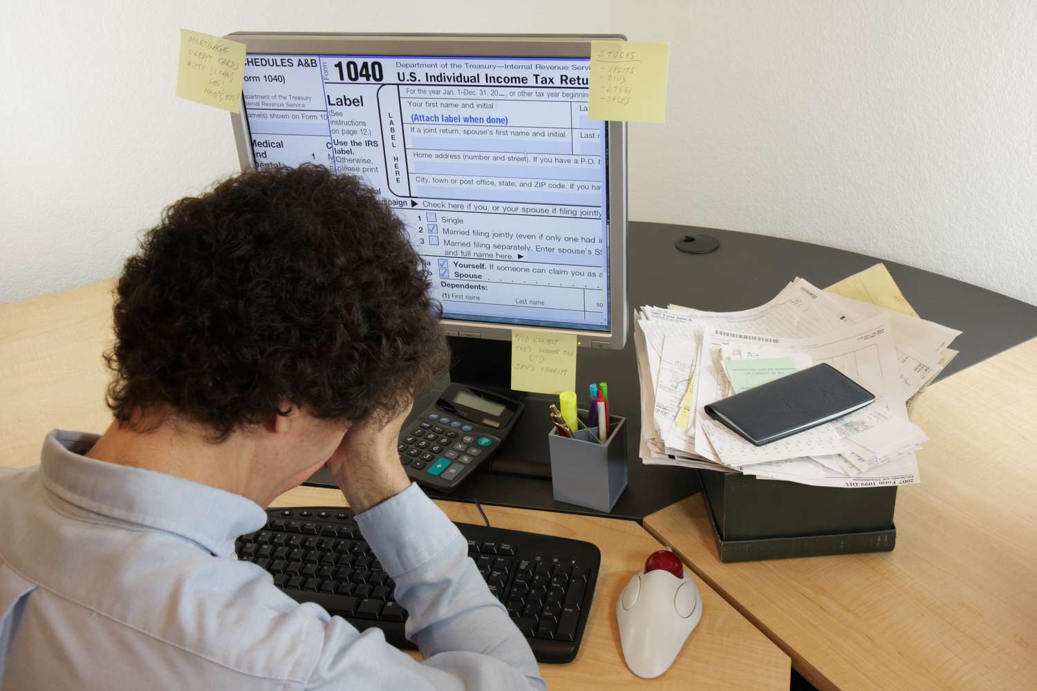A stressed taxpayer hold their head as they work on a desktop computer showing a IRS 1040 return with a nearby calculator, checkbook, and a stack of receipts