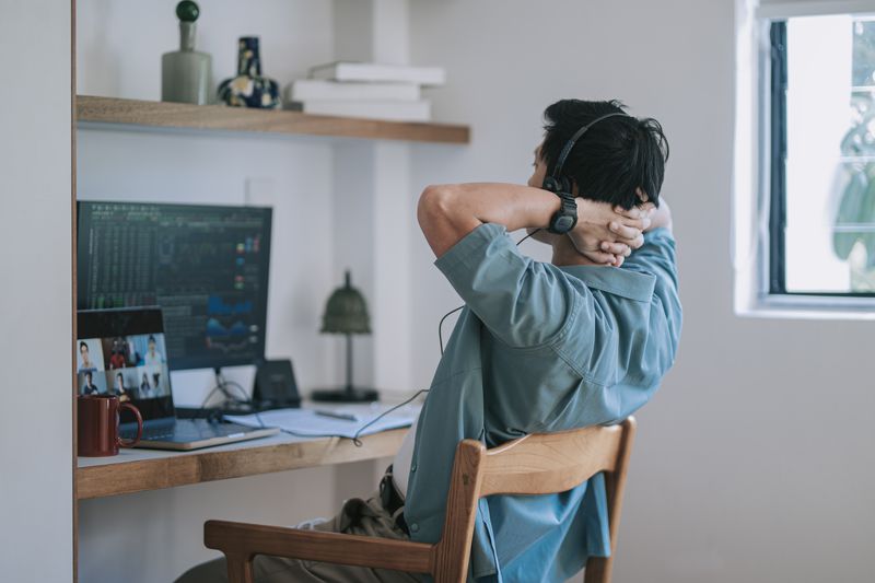 Man sitting at desk with hands crossed over back of head looking at monitor