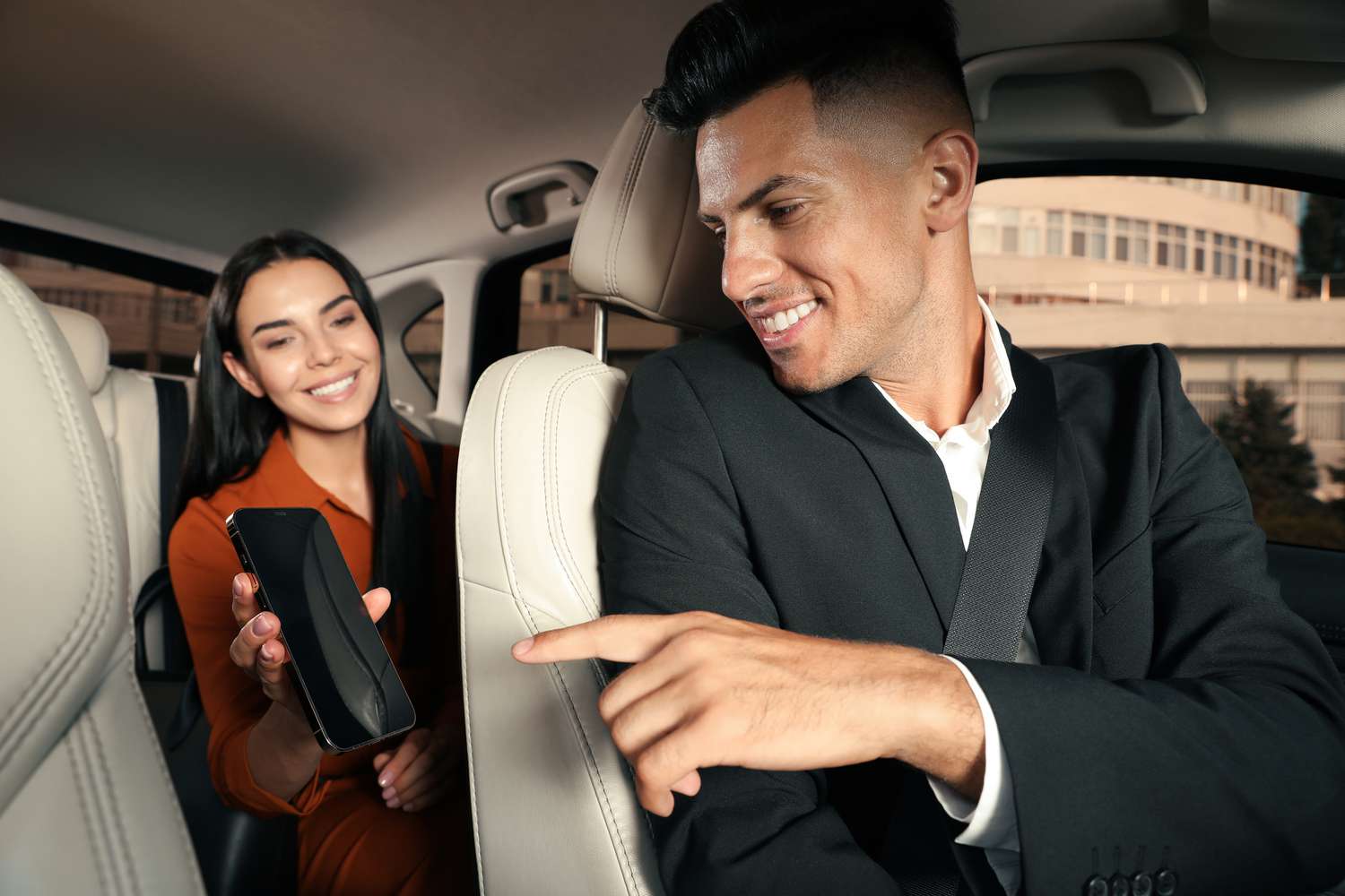 A man in a car suit pointing at a smiling woman holding a smartphone in the backseat