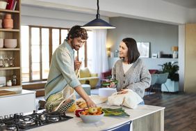 Happy couple in the kitchen taking out items from grocery bags and discuss about their reduced payments after a mortgage recast. 