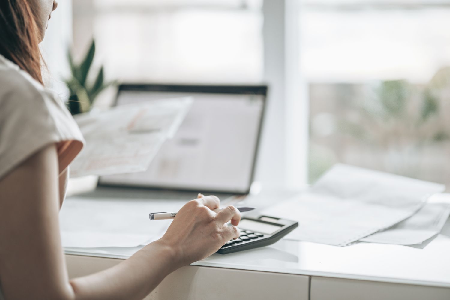 Woman typing on calculator at desk while holding papers, and a computer in the background