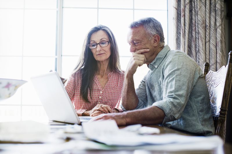 An older couple sits at a dining table, using a laptop computer with paperwork on the table