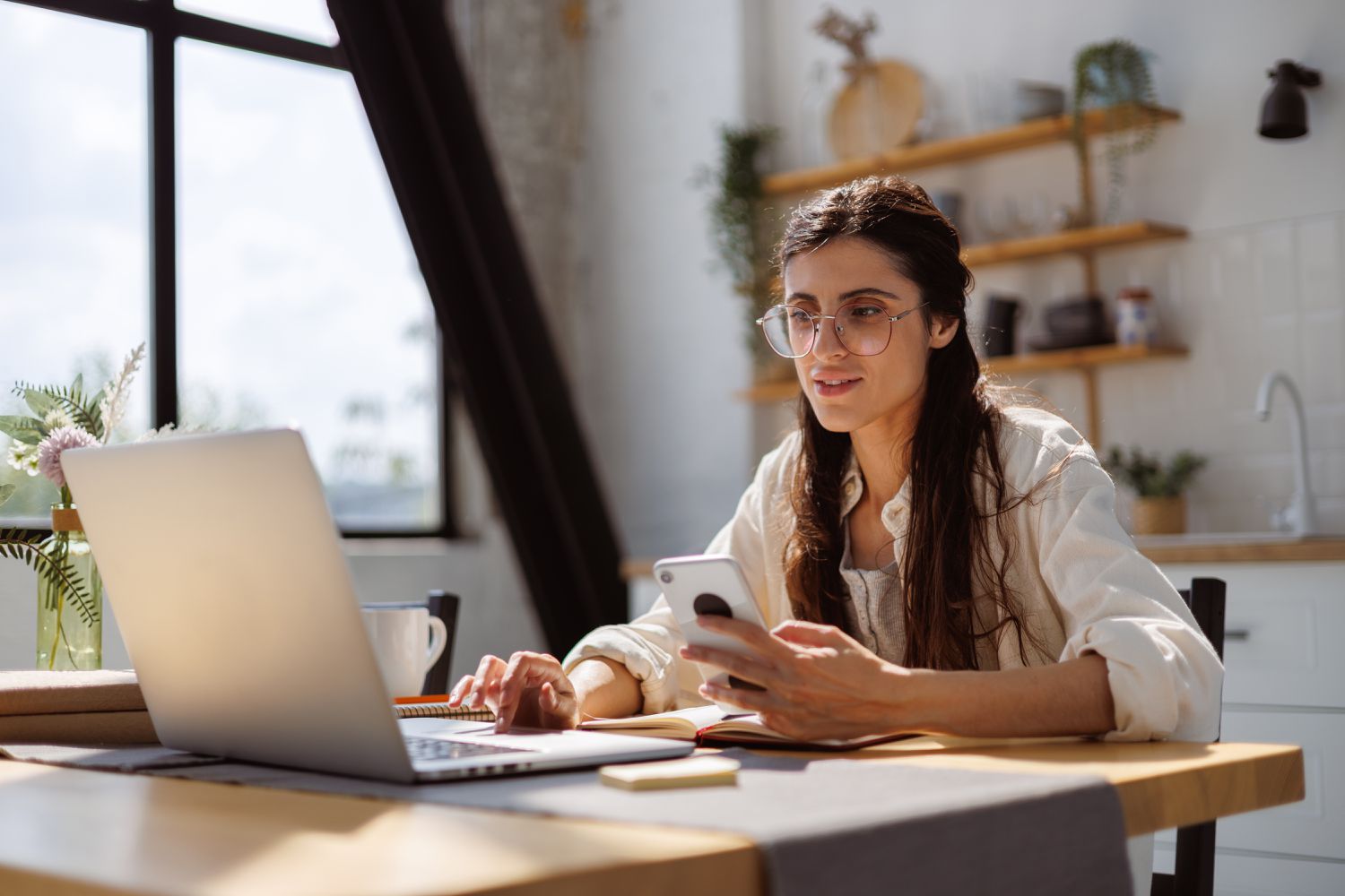 Young professional sitting in front of laptop with phone in hand looking into high-yield checking account benefits.