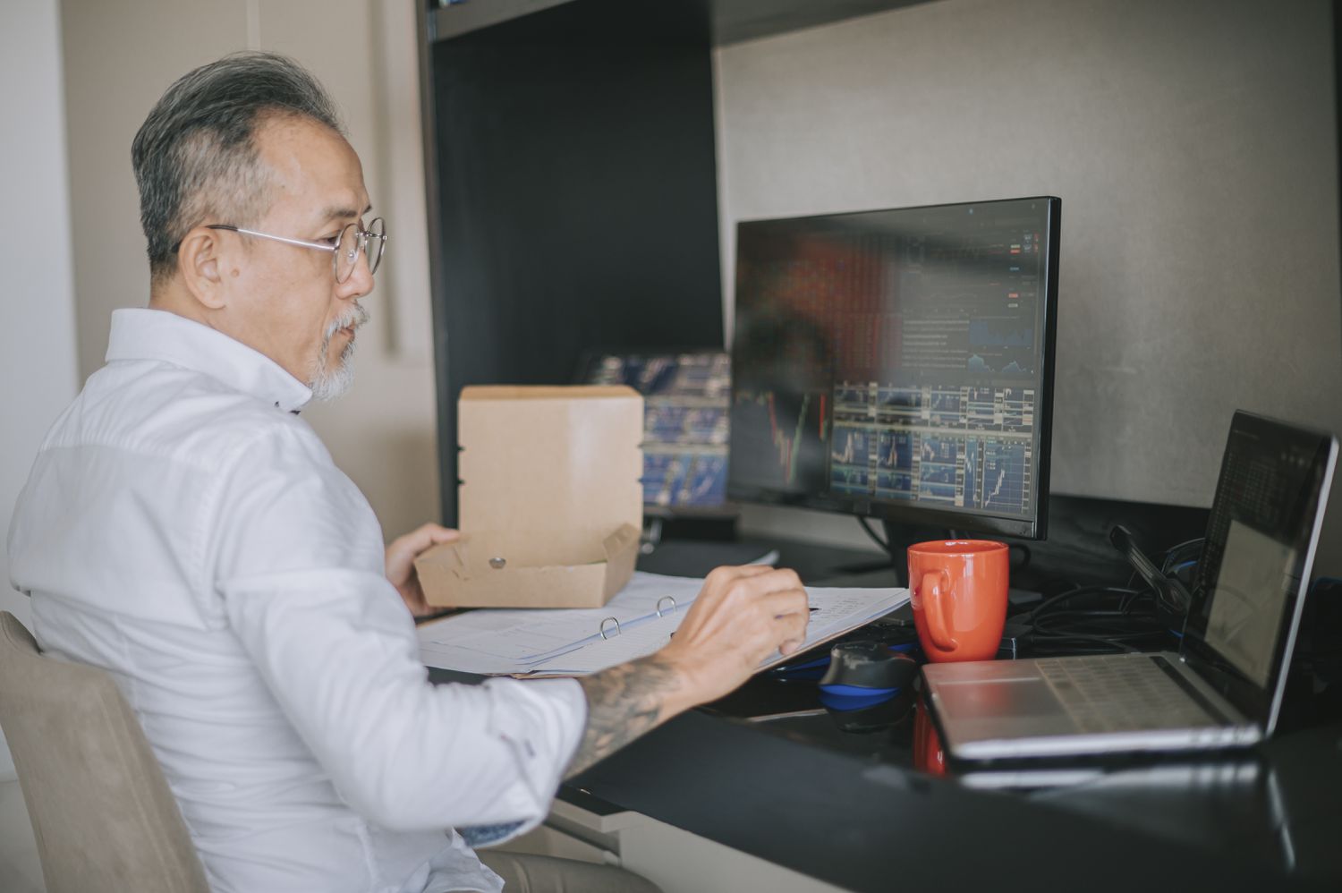 A retail trader sitting at him computer with an open laptop also on the desk