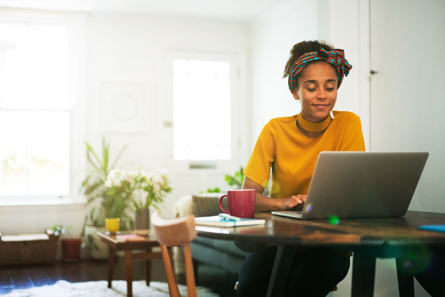 A woman in a yellow shirt sits at a table at home with a laptop and a red mug
