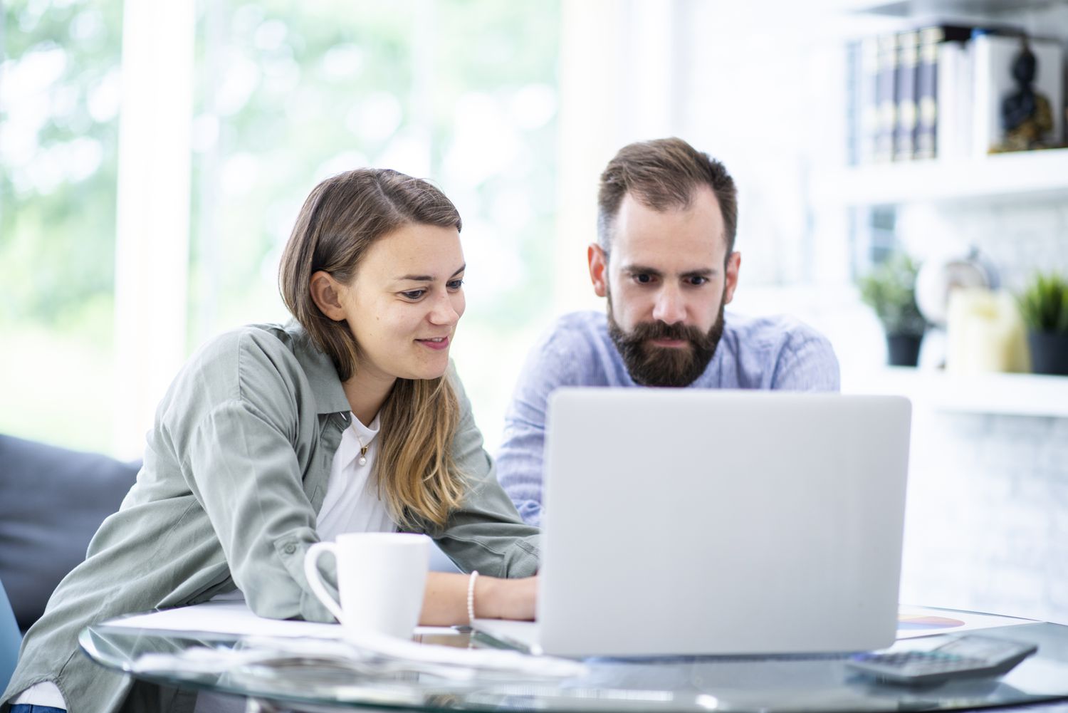 A couple in their 30s, sitting at their kitchen table, looks at a laptop screen together