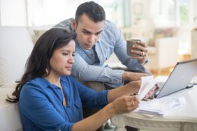 A man and a woman both wearing blue shirts sit at a coffee table with papers and a laptop; the man is holding a mug