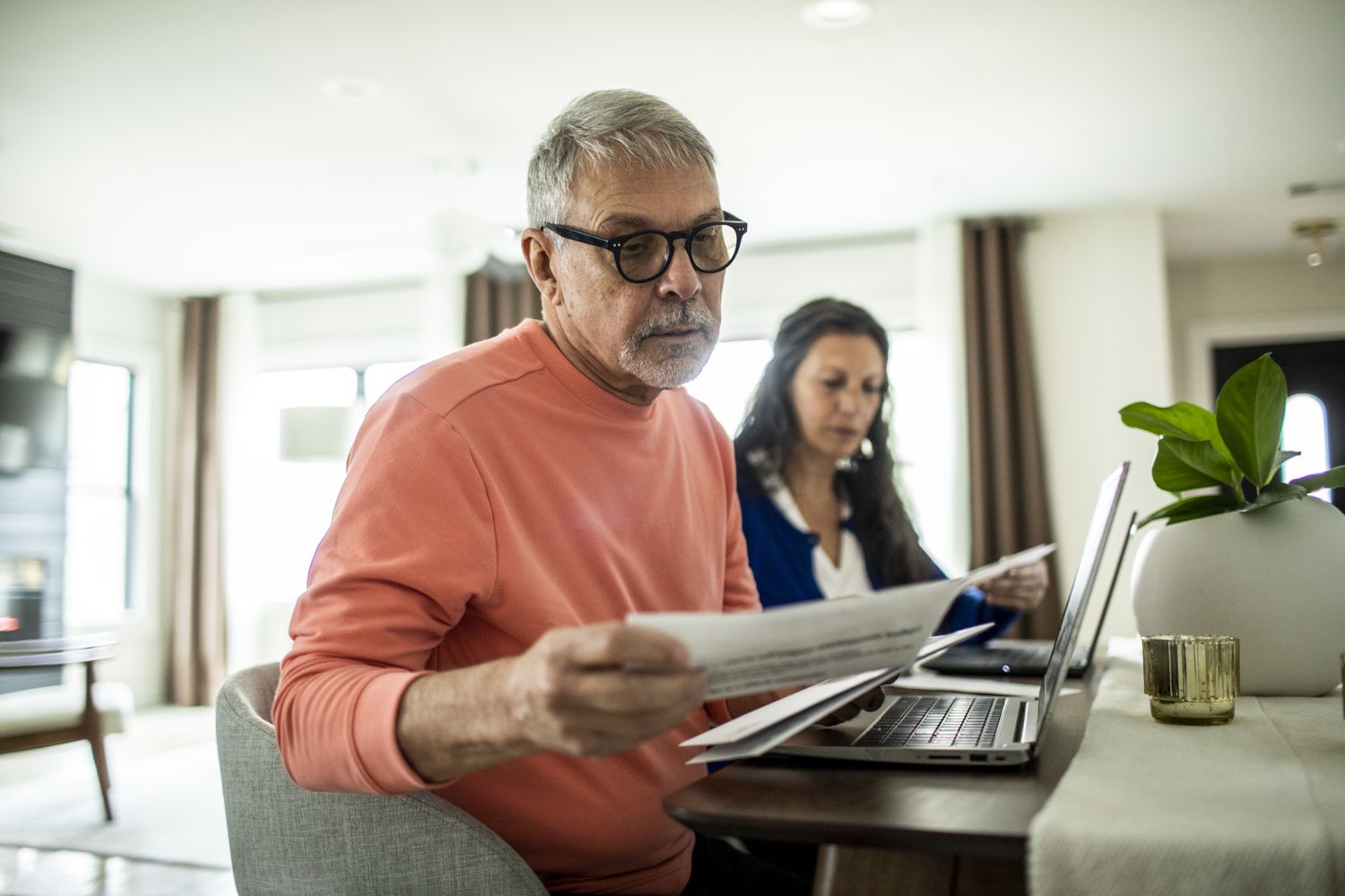 A man and a woman sit at a desk or table and review documents. In front of each is an open laptop.
