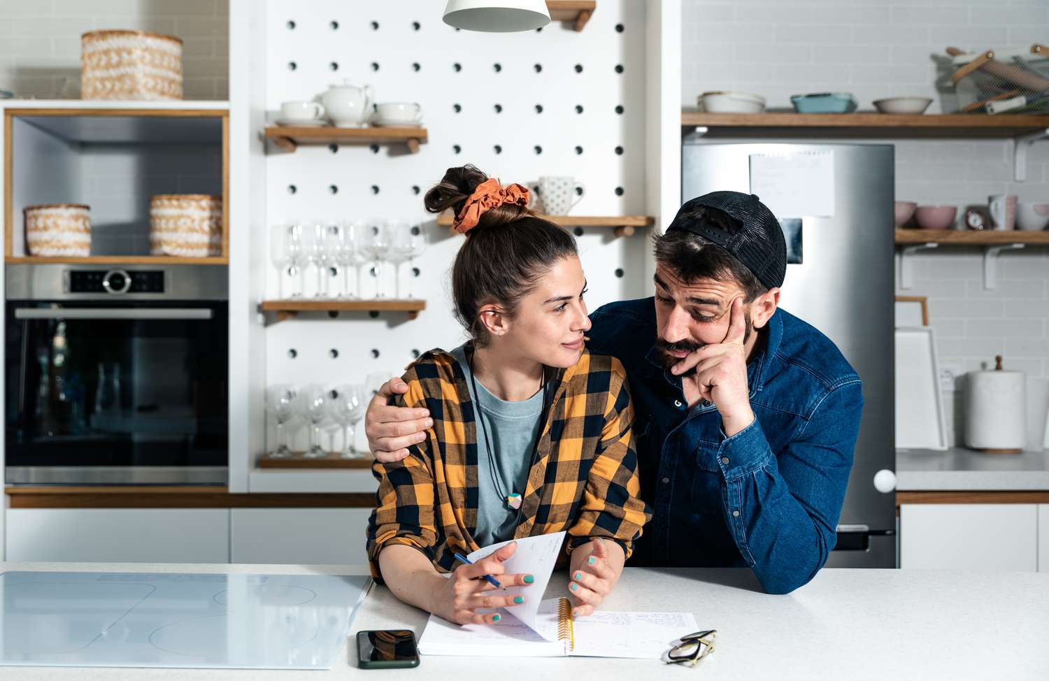 A man and a woman stand against a kitchen island discussing something. There's a notebook, phone, and glasses on the counter.