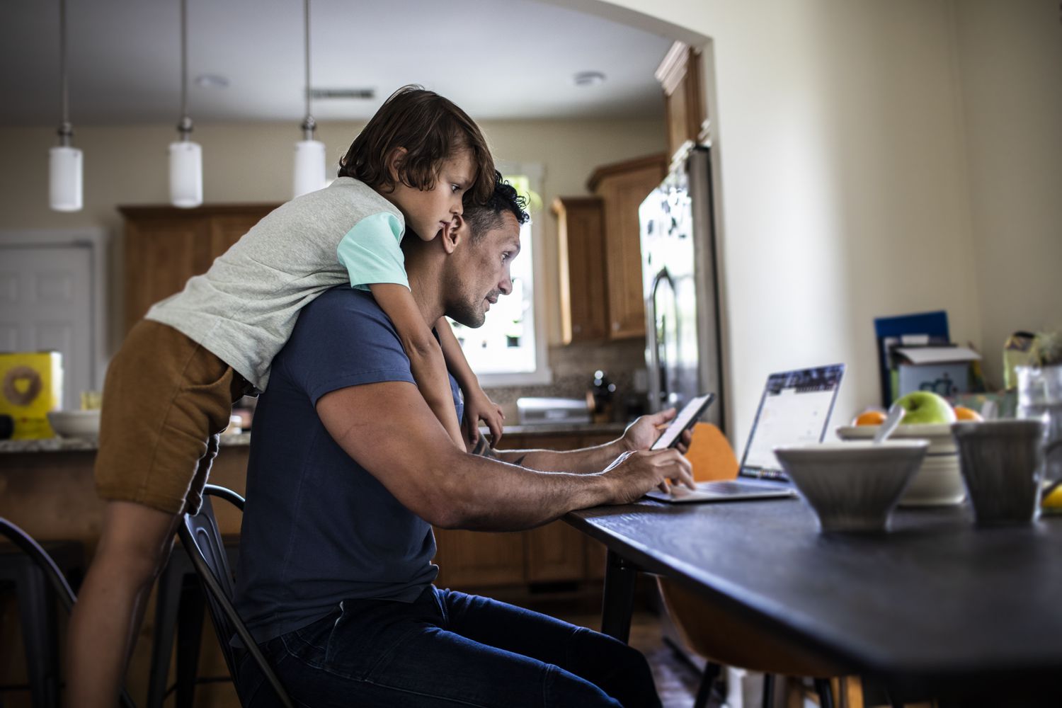 Father using laptop at table in kitchen while son looks over his shoulder