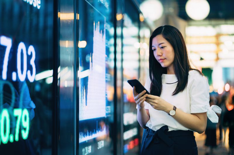 Young Asian businesswoman checking financial trading data on smartphone by the stock exchange market display screen board in a downtown financial district
