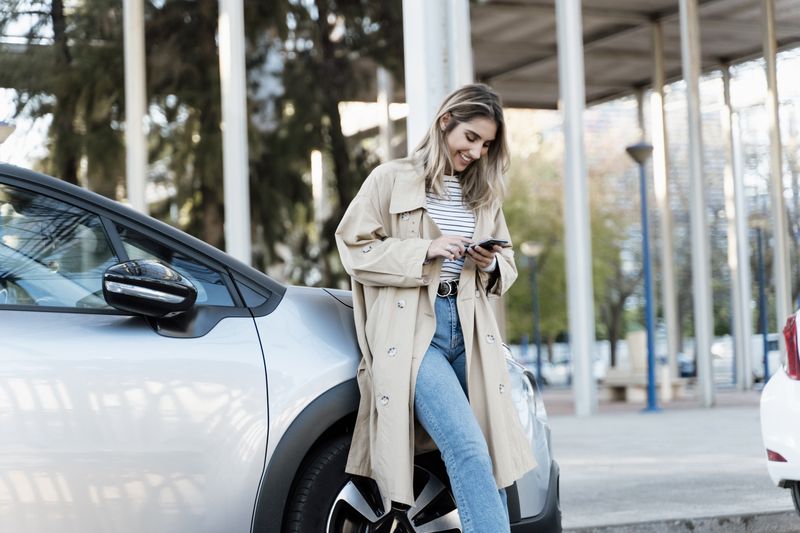 A woman leans on a car and uses a phone.